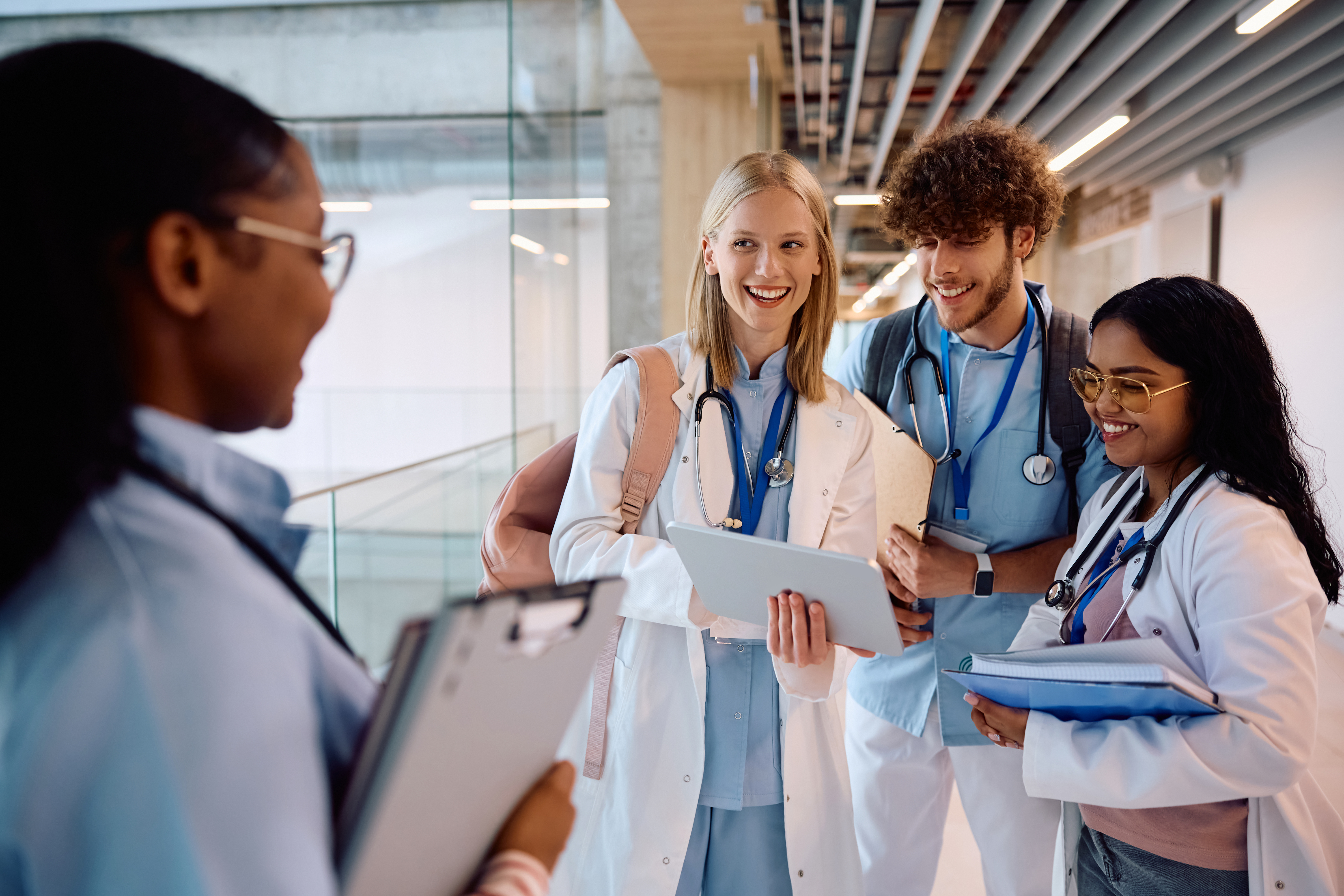 Eine Gruppe Medizinstudierende steht auf dem Gang in einer Universität und begrüßt freundlich lachend eine weitere Studentin, die auf sie zugelaufen kommt.